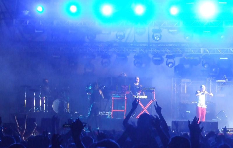 The Prodigy perform on a festival stage in Romania under blue lights, with a crowd's raised hands in the foreground.
