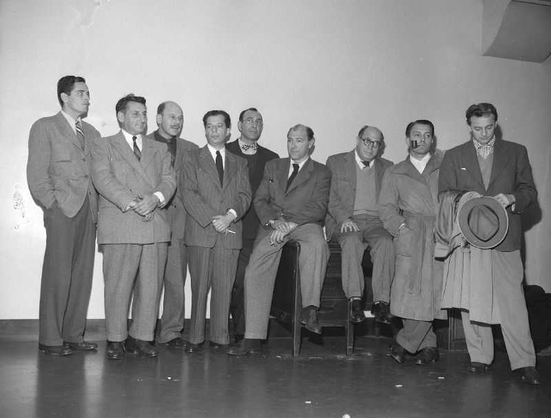 Nine men in suits and overcoats stand and sit in a row against a bare wall, one holding a hat