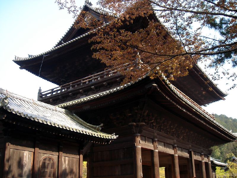 A dark wooden temple gate with tiered roofs beside autumn trees.