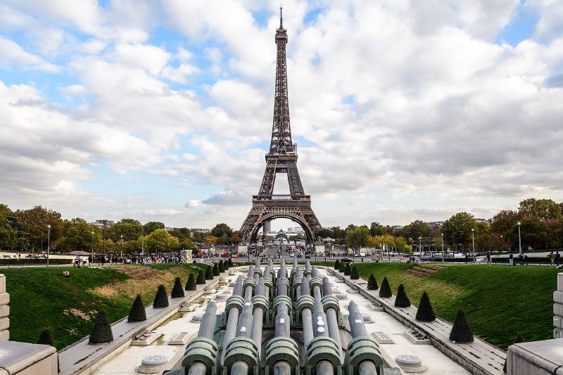 Colour photograph of the Eiffel Tower seen along the same axis from the Trocadéro, with fountain jets in the foreground and a blue, cloudy sky.