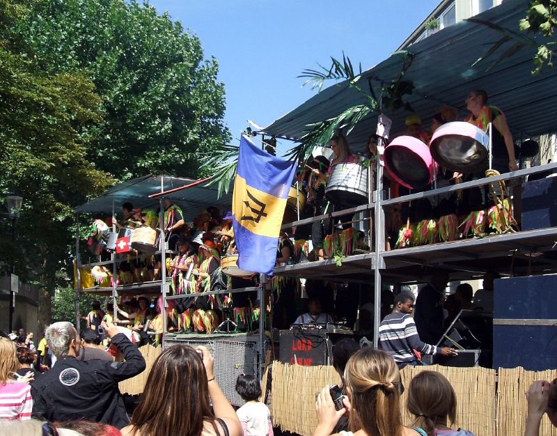 A Notting Hill Carnival float carries steel pan players above a DJ's turntables and a stack of speakers, with a Barbados flag and a crowd below.