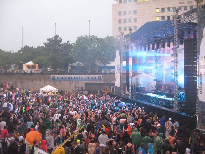 A crowd with umbrellas fills an outdoor festival site in front of a lit stage.