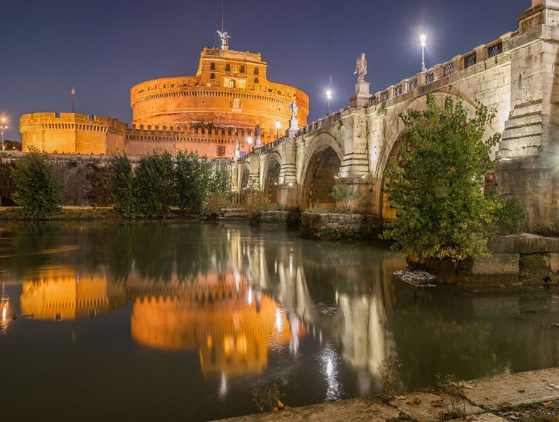 Castel Sant'Angelo and its stone bridge lit at night, reflected in the River Tiber in Rome.