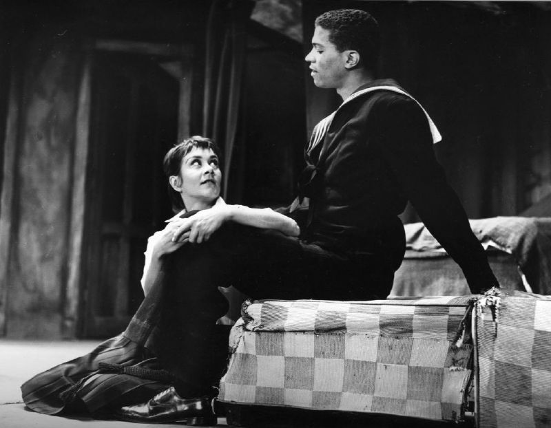 A black-and-white stage photograph of a young woman in a school blouse sitting on the floor, looking up at a young man in a sailor's uniform seated on a chequered bed.