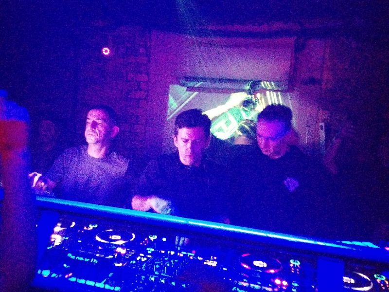 Three drum and bass DJs stand behind CDJ decks in a dark, blue-lit London club booth.