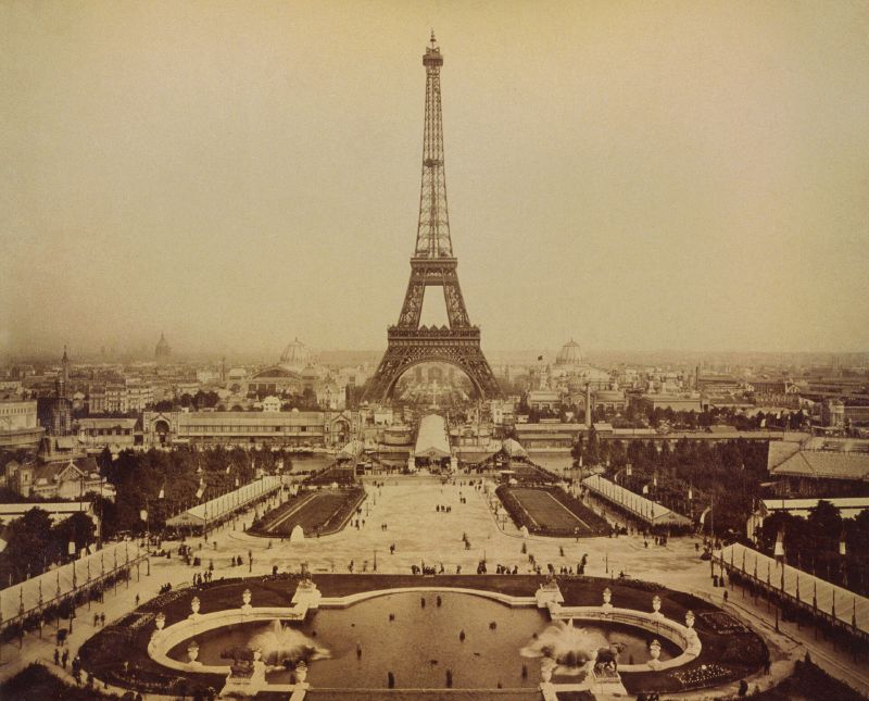 Sepia photograph of the Eiffel Tower seen from the Trocadéro, with a fountain basin in the foreground and the Champ de Mars exhibition grounds beyond.