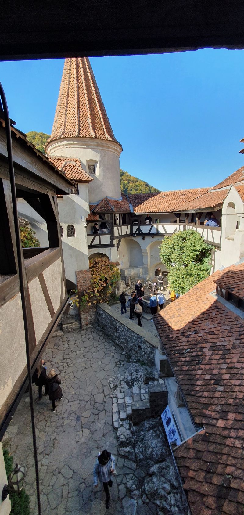 A stone courtyard seen from above, with a tall red-tiled round tower, timber balconies and visitors below.