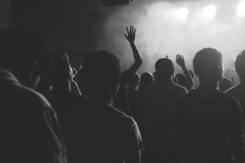 The backs of a dark concert crowd, with raised hands and stage smoke lit from behind.
