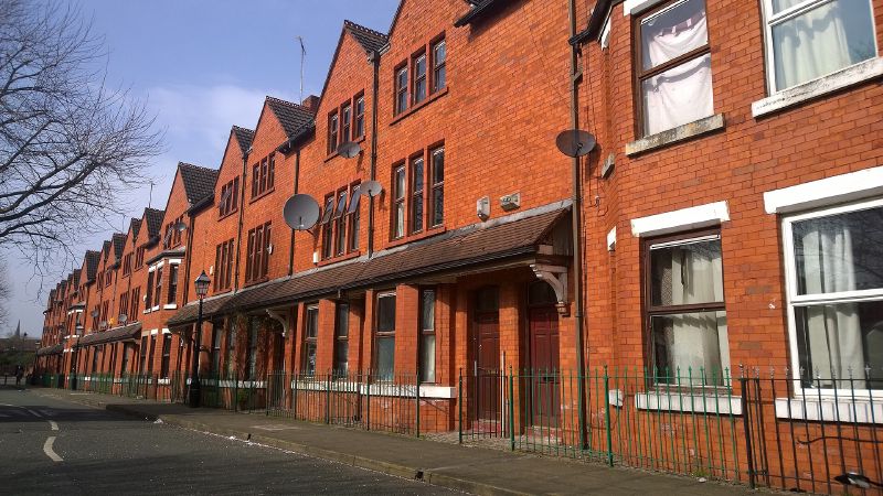 A row of red-brick terraced houses with satellite dishes, green railings, and a tiled canopy over the front doors, under a blue sky.