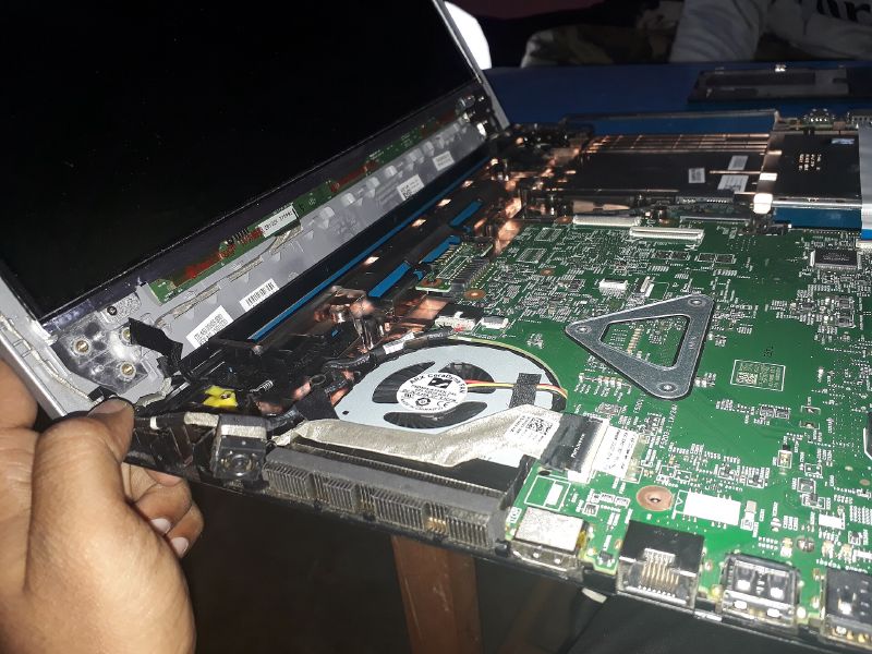 A laptop opened up on a desk, with the screen lifted away to show the green motherboard, the cooling fan and memory slots, held steady by a person's hand.