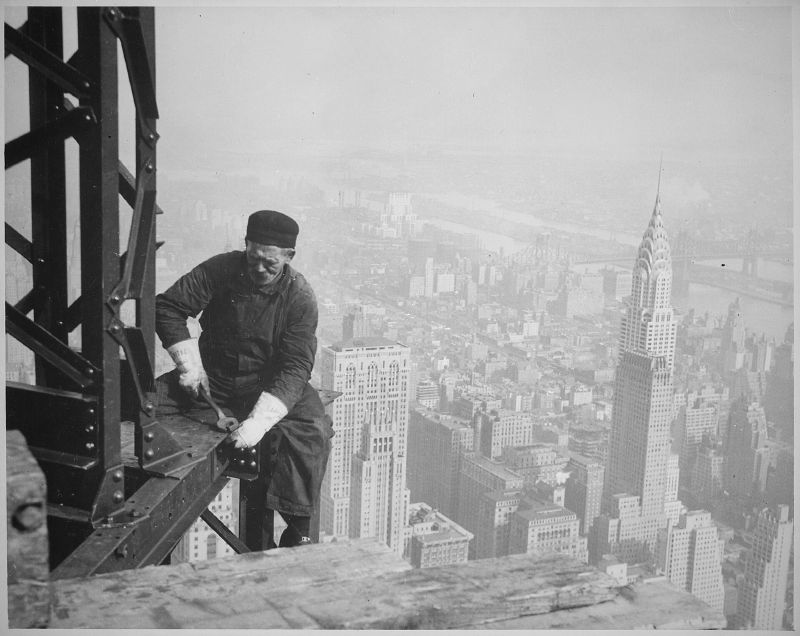 An older ironworker in a cap and gloves sits on a steel beam high on the Empire State Building's frame, the Chrysler Building's spire behind him