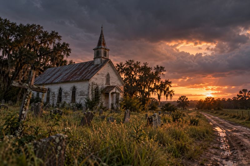 A weathered white country church and overgrown graveyard under a storm sky at sunset.