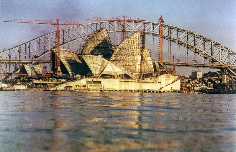 The Sydney Opera House under construction, its tiled shells nearly complete beside a crane, seen across the water from Farm Cove