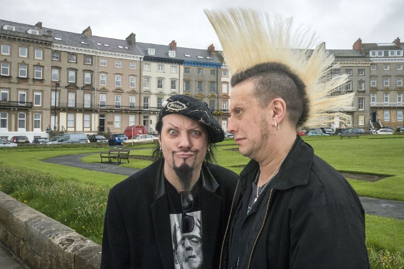 Two men in black clothes pose in front of a grassy square and terraced buildings, one with a tall bleached mohican.
